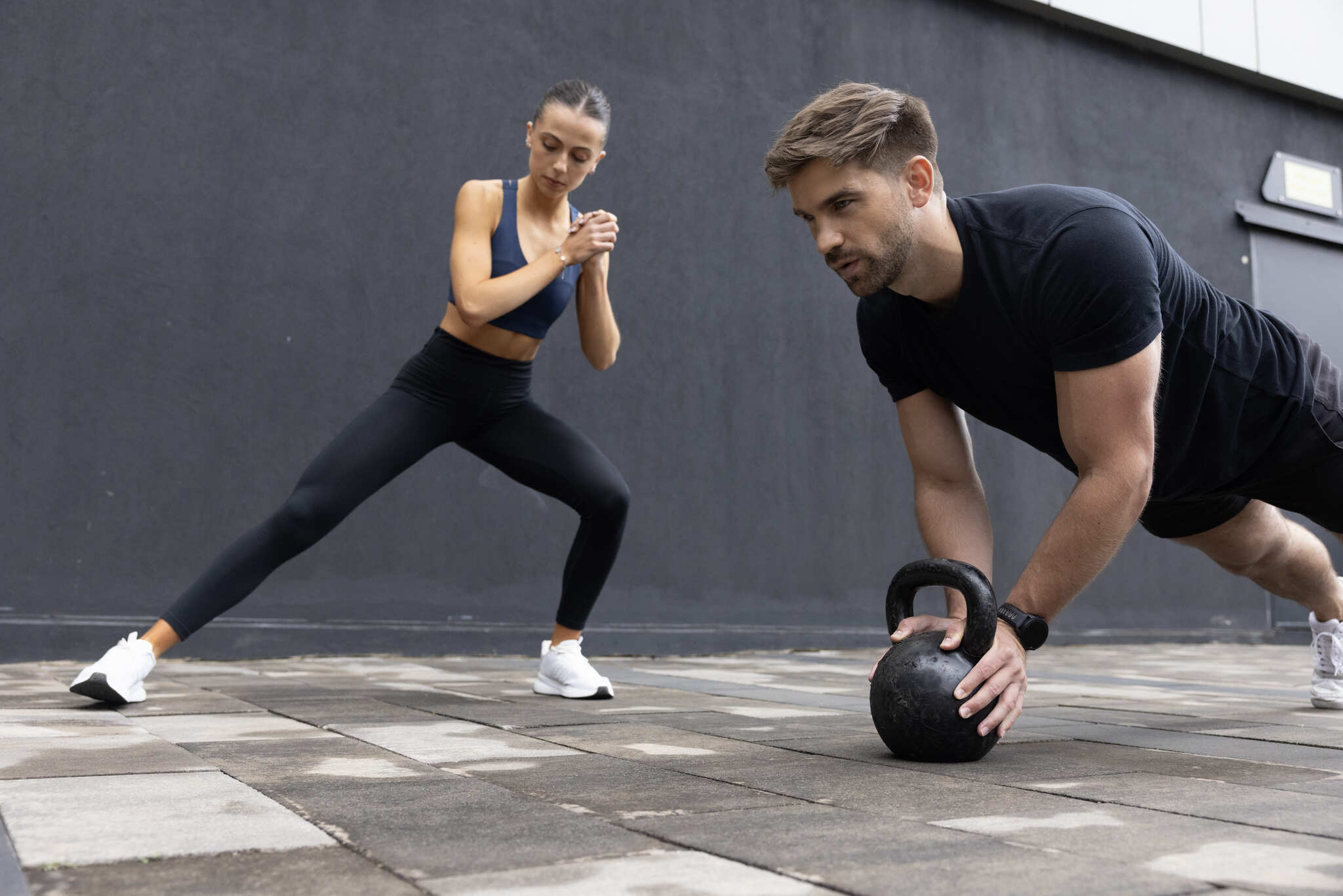 Two people exercising outdoors. A man is in a plank position holding a kettlebell, and a woman is doing a side lunge.